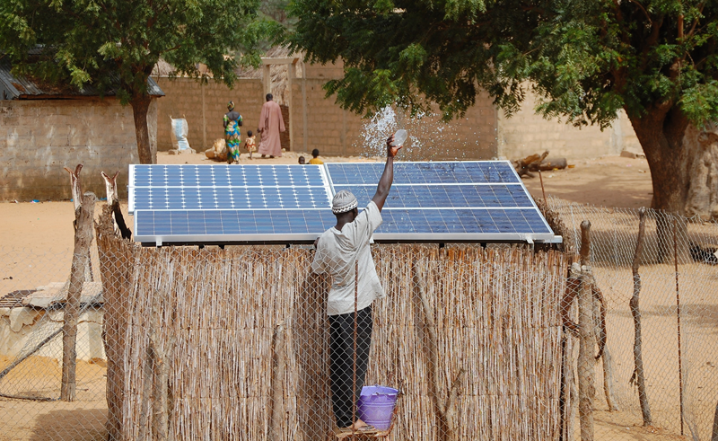 Hombre subido a un banco y limpiando las placas solares de su casa de paja ubicada en África.